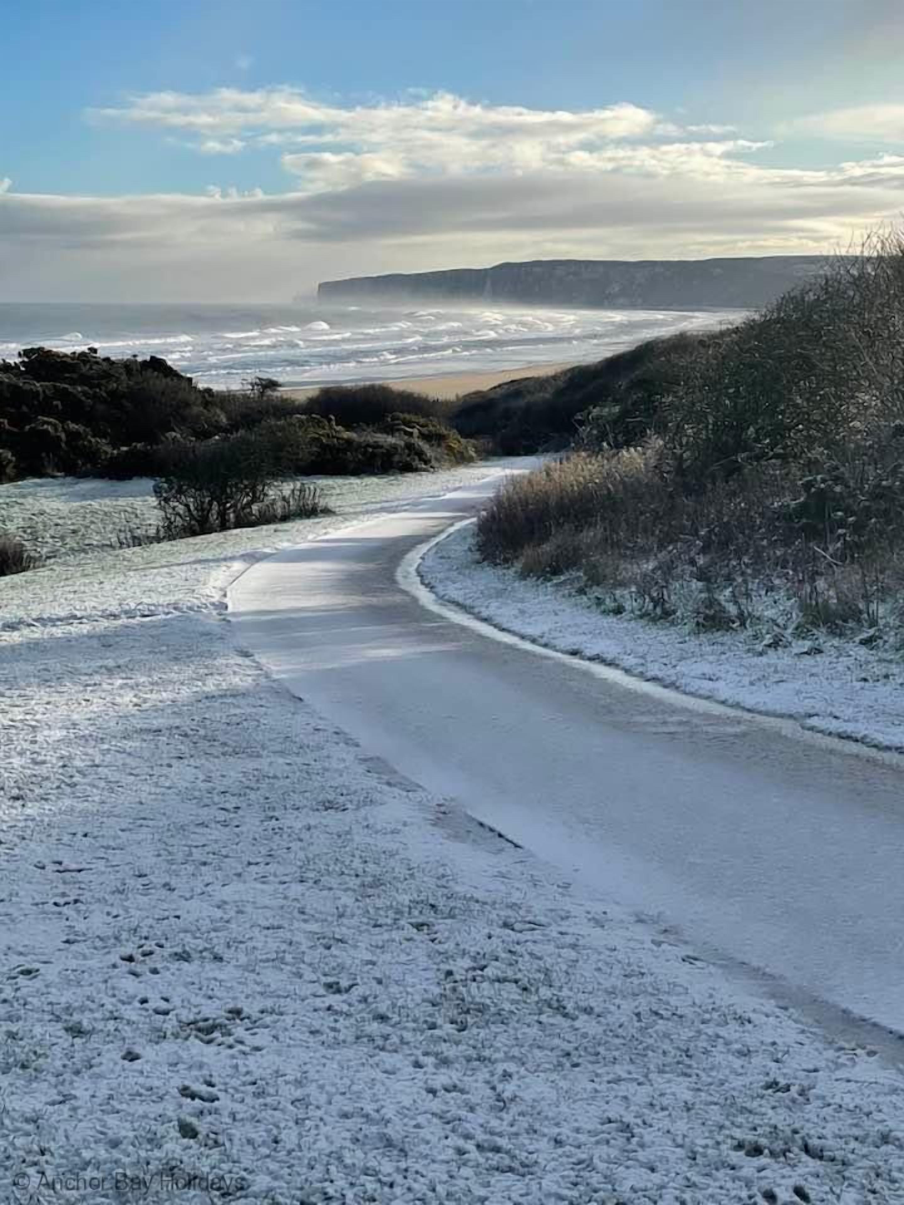 Eine verschneite Straße schlängelt sich durch eine natürliche Landschaft, im Hintergrund Meer und Klippen.