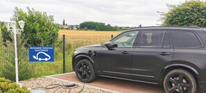 Un SUV gris estacionado en un área de grava con una estación de carga, rodeado de follaje y un campo.