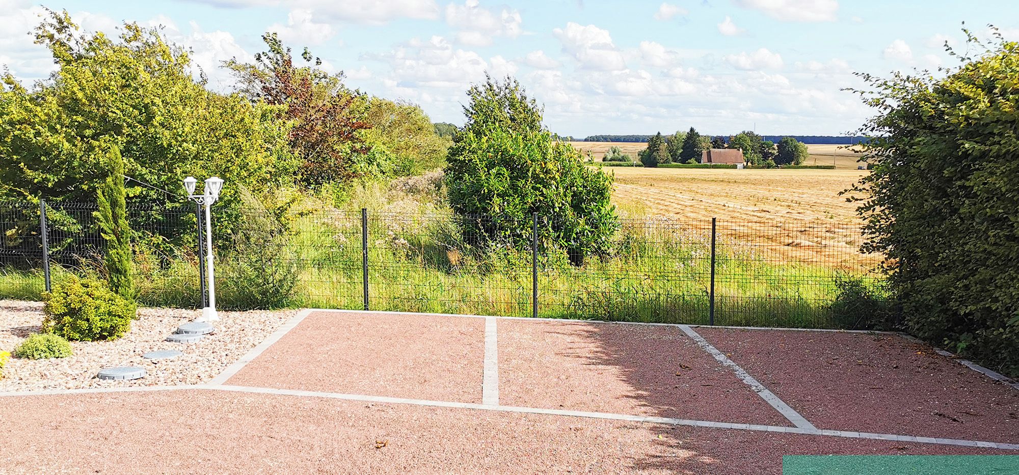 A tennis court with a fence, trees, and a field in the background.