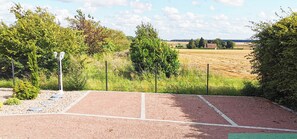 Una cancha de tenis con valla, árboles y un campo al fondo.