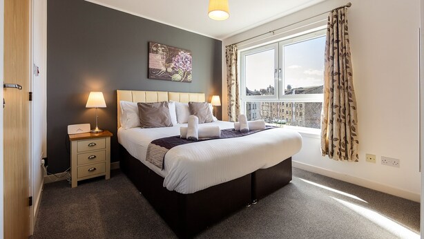 A neatly arranged bedroom with a large bed, bedside lamps, a nightstand, and a window with curtains. - Modern, Spacious City Apartment, Lift, Balcony, Free Wi-fi & Parking, Fab views, (Edinburgh)