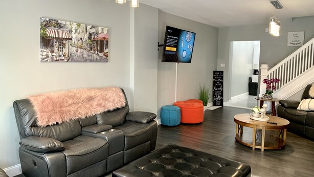 Ett modernt vardagsrum med en svart lädersoffa, en stoppad pall och ett soffbord. En platt-TV är monterad på väggen, en trappa syns i bakgrunden och ovanför soffan hänger en inramad tavla.