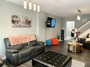 Un salon moderne avec un canapé en cuir noir, un pouf capitonné et une table basse. Un téléviseur à écran plat est fixé au mur, un escalier se trouve à l’arrière-plan et un tableau encadré est accroché au-dessus du canapé.