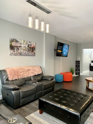 A modern living room with a black leather sofa, a tufted ottoman, and a flat-screen TV mounted on the wall.