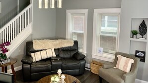 Ett vardagsrum med en svart lädersoffa, en beige fåtölj, ett träbord och en trappa med vita räcken.
