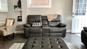 Ett modernt vardagsrum med en svart lädersoffa, en stoppad pall och en beige fåtölj. Det finns en dekorativ vägghylla med en växt och en skylt, ett fönster med ett dekorativt galler och en vit dörr.