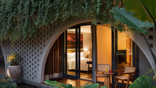 Una habitación de hotel moderna con un amplio ventanal corredero que da a un balcón, una zona de estar con mesa y sillas, y una planta en maceta.