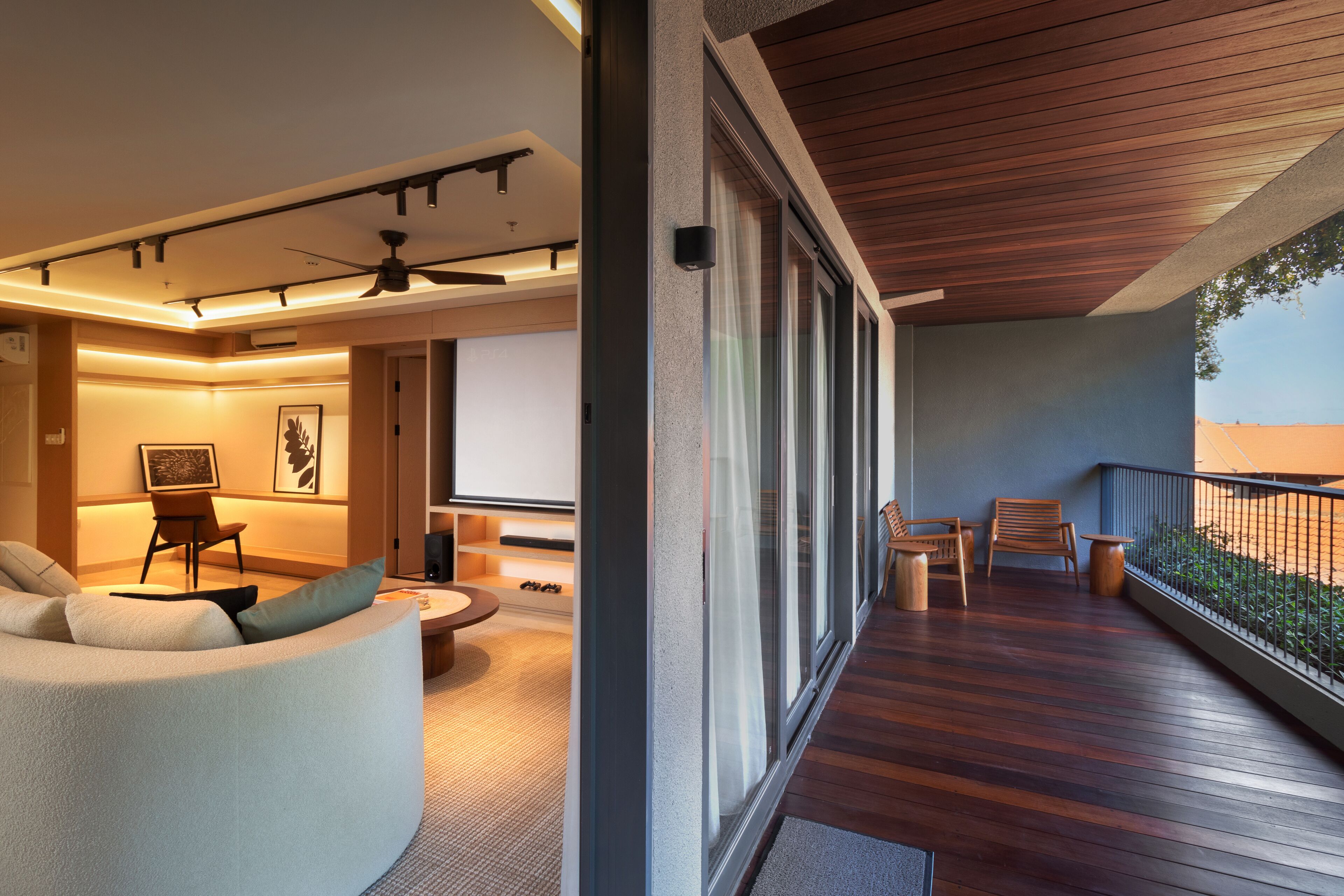 A modern living room with a curved sofa, a coffee table, and a TV. There is a balcony with wooden flooring and a railing.