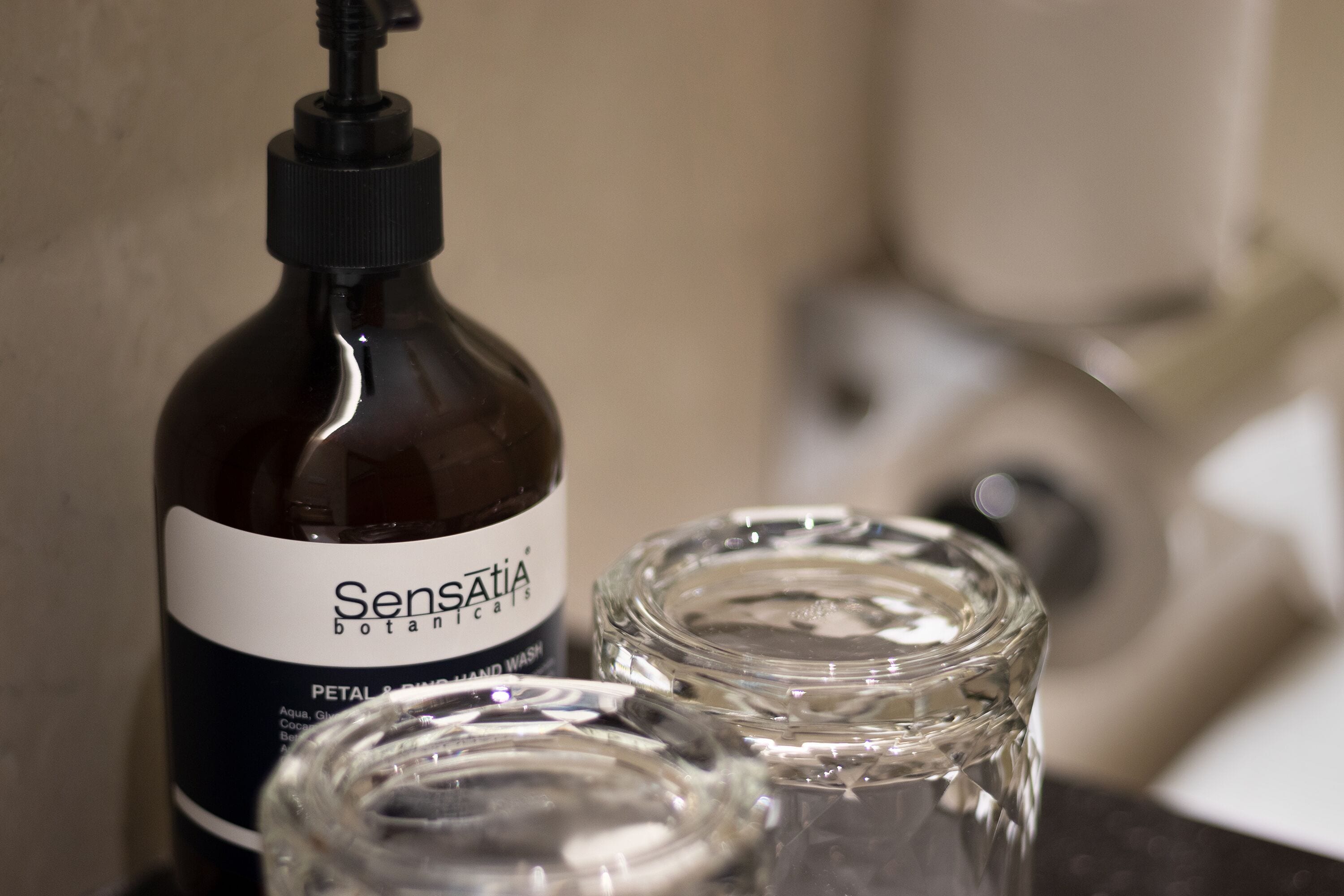 Una botella de jabón de manos Sensatia Botanicals junto a dos recipientes de vidrio transparente.