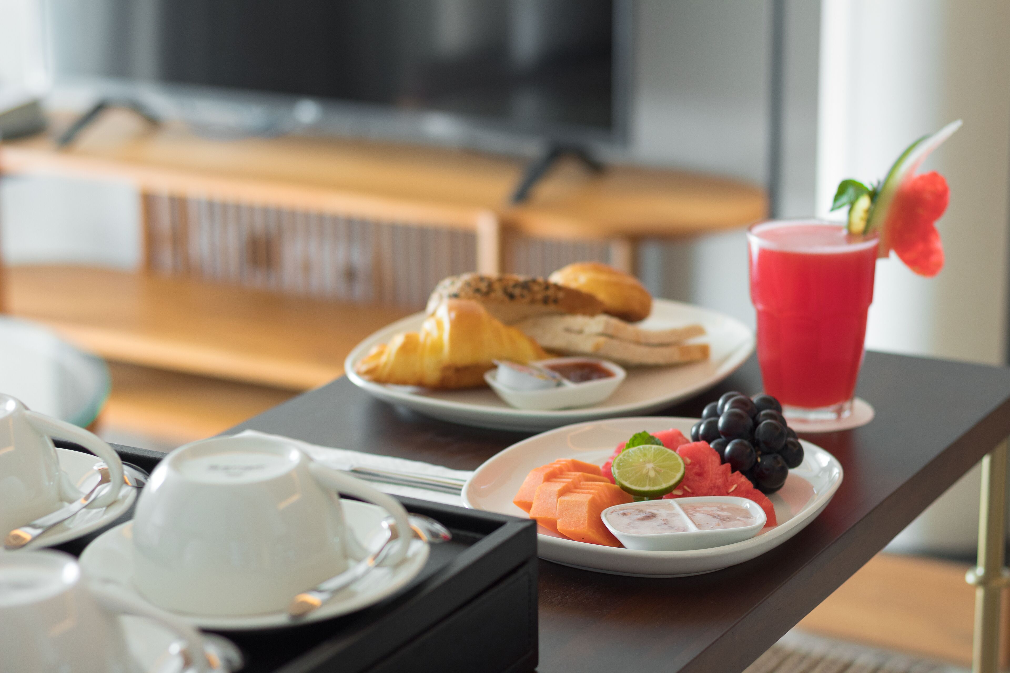 Een ontbijtkarretje met croissants, een fruitschaal met watermeloen, druiven en een limoen, en een rood drankje met garnering.