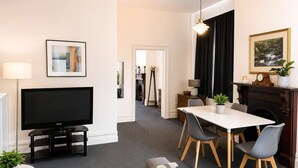 Ein modernes Wohnzimmer mit Flachbildfernseher, einem Esstisch mit Stühlen, einem Kamin und einem Flur, der zur Küche führt.