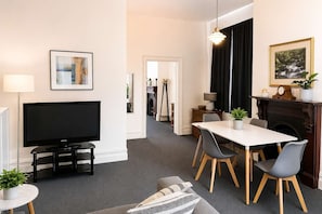 A modern living room with a flat-screen TV, dining table, chairs, a sofa, a fireplace, and a hallway leading to a kitchen.