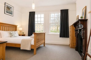 A hotel room with a wooden bed, a nightstand, a lamp, a fireplace, and a view of a building through two windows.