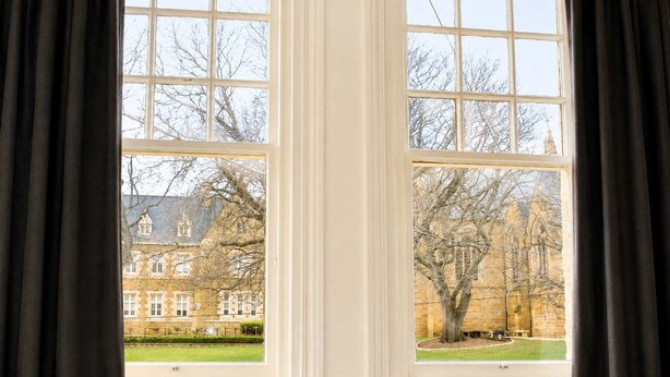 Ein Zimmer mit zwei großen Fenstern, Blick auf ein Gebäude mit Ziegelaußenwand und einem Rasen.