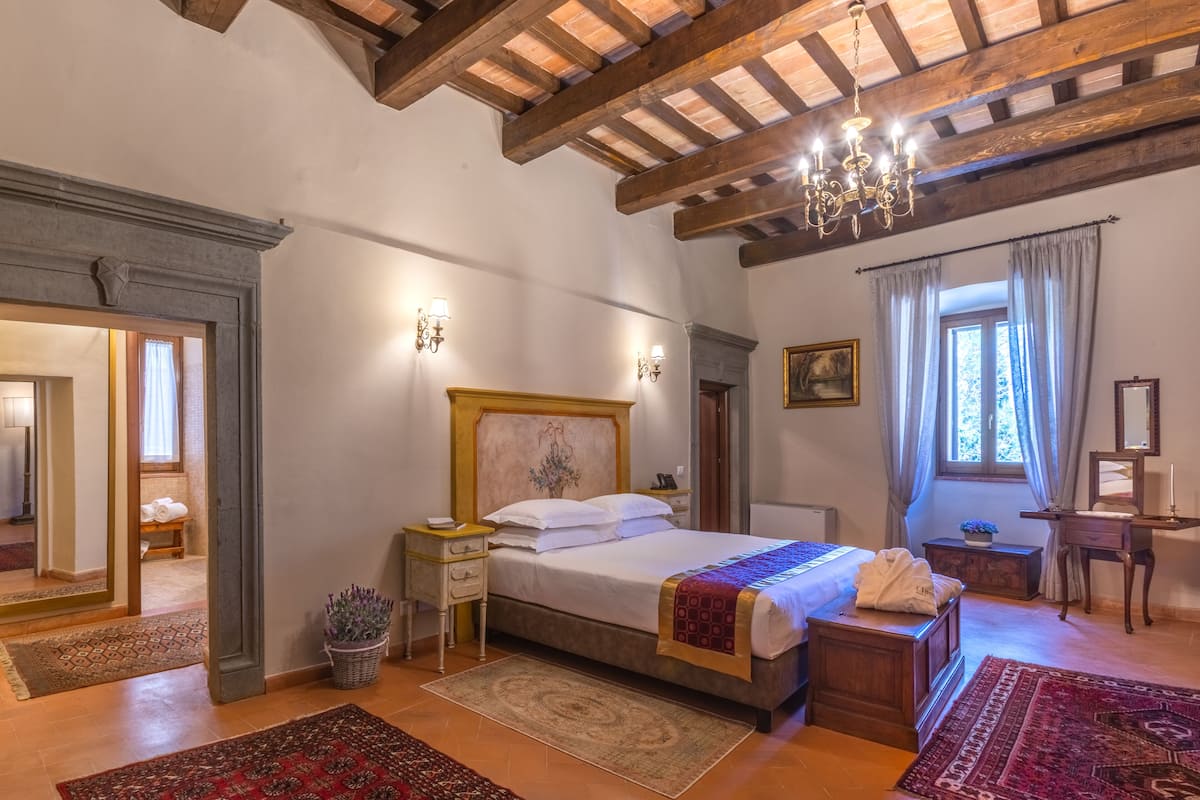 Un dormitorio con techo de madera, una cama con cabecera, una mesita de noche, un aparador, un espejo, una ventana con cortinas y una planta en maceta.