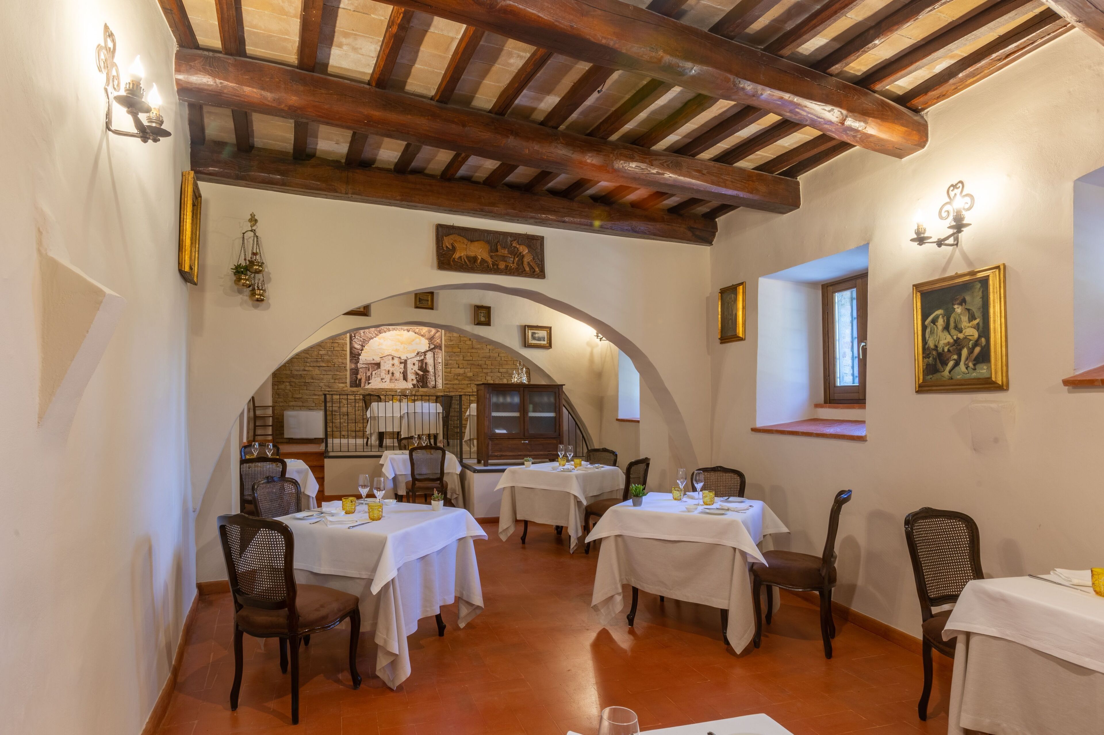 Un espace repas avec des tables disposées pour accueillir quatre personnes, des poutres en bois au plafond et un mur en briques agrémenté d’une fenêtre.