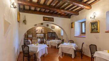 Un espace repas avec des tables disposées pour accueillir quatre personnes, des poutres en bois au plafond et un mur en briques agrémenté d’une fenêtre.