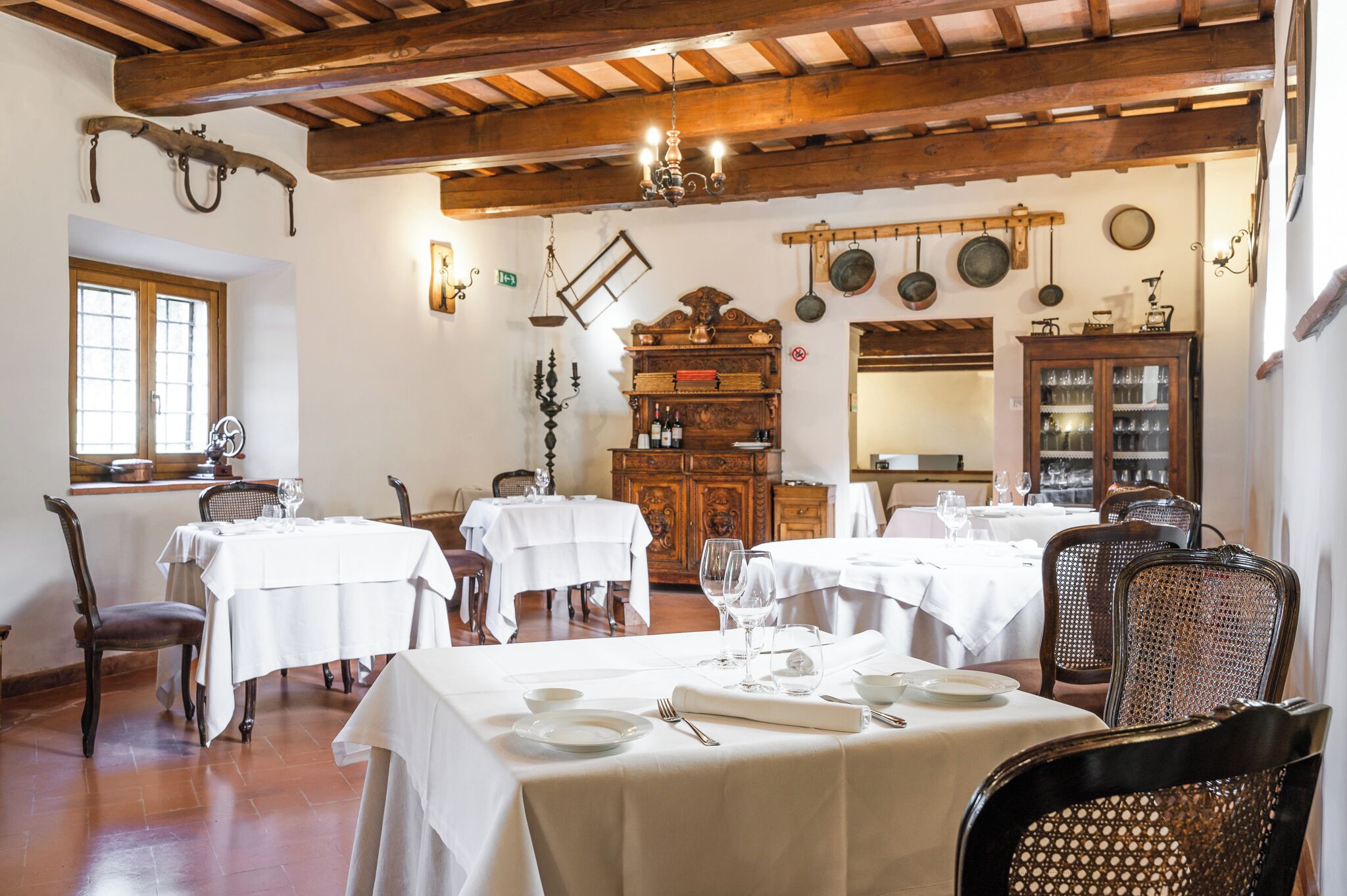 A dining area with wooden beams, white tables, and chairs. There are hanging pots and a wooden cabinet with glassware.
