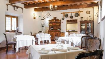A dining area with wooden beams, white tables, and chairs. There are hanging pots and a wooden cabinet with glassware.