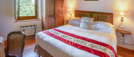 A bedroom with a large bed, wooden beams on the ceiling, a window with a view of trees, and a wooden wardrobe.