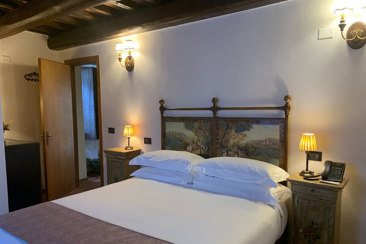 Una cama bien hecha con sábanas blancas, dos lámparas de noche, un cabecero de madera con un cuadro y un teléfono sobre una mesita de noche.