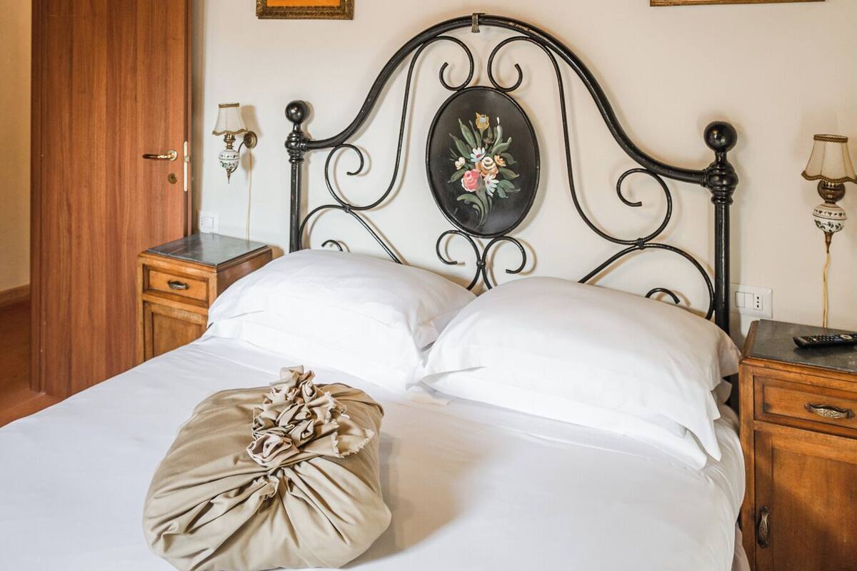 Un dormitorio con techo de madera, una cama con ropa de cama blanca, un cojín decorativo, una mesita de noche con una lámpara y un cuadro en la pared.