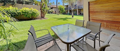 An outdoor patio with a glass table and metal chairs, surrounded by a well-maintained lawn and palm trees.