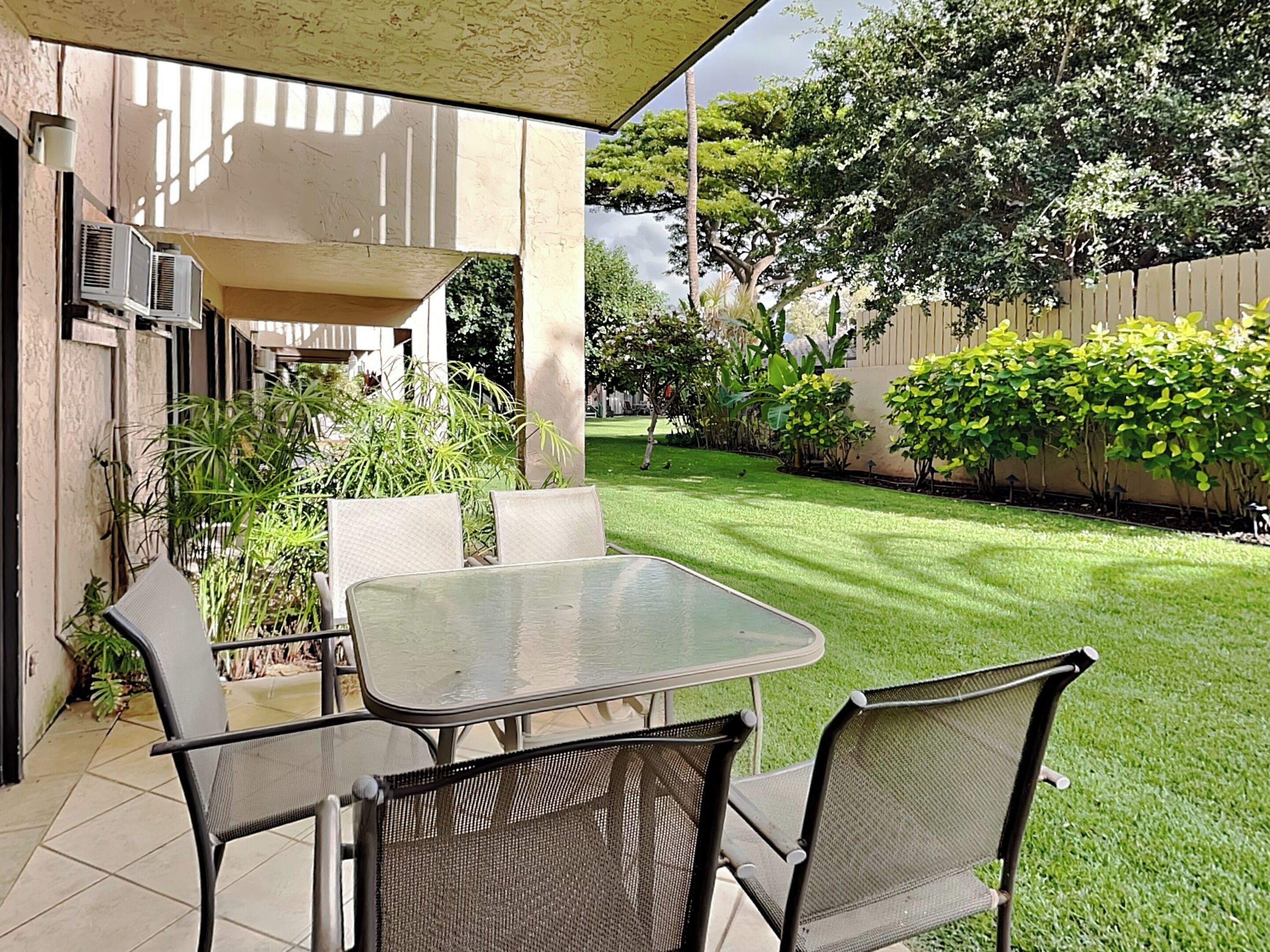 An outdoor patio with a glass table and metal chairs, surrounded by a well-maintained lawn and various plants.