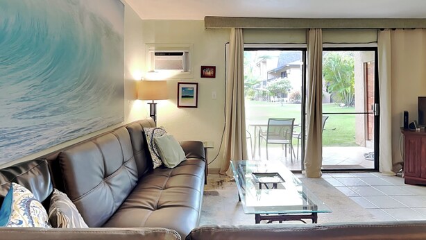Un salon comprenant un canapé en cuir, une table basse en verre, une grande fresque murale, un ventilateur de plafond et une porte-fenêtre coulissante donnant sur un espace extérieur.