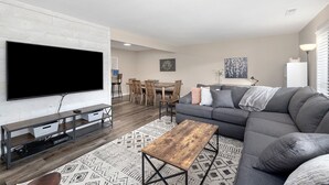 A modern living room with a flat-screen TV, a gray sofa, a wooden coffee table, and a dining area with wooden chairs and a table. - WALK IN, Pointe Royale, Arcade, Indoor Pool (Branson)