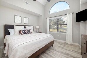 A bedroom with a large bed, a window with blinds, a flat-screen TV, and a small bedside table.