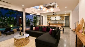 A modern living room with a black sectional sofa, a round coffee table, and a dining area with a kitchen in the background. - Villa Soho (Canggu)