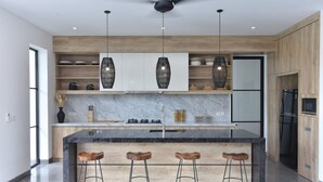 A modern kitchen with a marble countertop, wooden bar stools, pendant lights, and built-in shelving. - Villa Soho (Canggu)