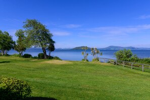 A grassy area with a wooden fence, trees, and a body of water with distant hills.