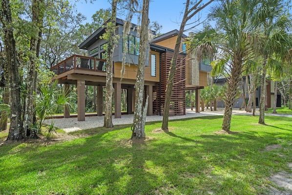 A modern two-story house with a balcony, surrounded by lush greenery and trees.