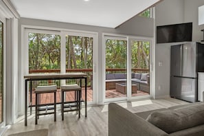 A modern kitchen with a built-in refrigerator, a dining area with a table and chairs, and a large sliding glass door leading to an outdoor deck with a view of a lush garden.