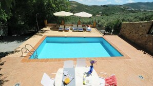 Ein Schwimmbecken mit Liegestühlen und Sonnenschirmen, umgeben von einer gefliesten Terrasse und Blick auf Hügel.