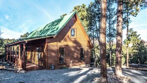 A log cabin with a green roof, a porch, and a gravel driveway. - Wine N Pines by Ruidoso Vacation Rentals (Alto)