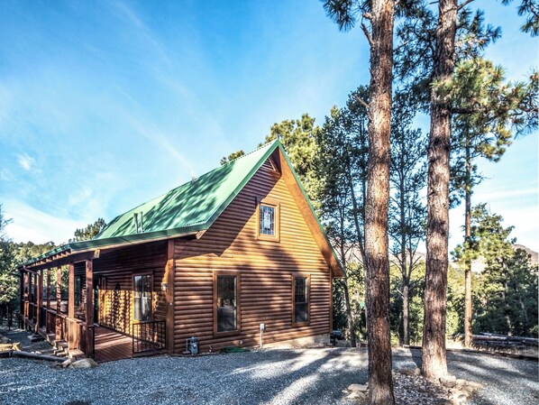 A log cabin with a green roof, a porch, and a gravel driveway. - Wine N Pines by Ruidoso Vacation Rentals (Alto)