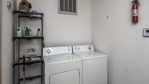 A laundry room with two white front-loading washers and dryers, a metal shelf with cleaning supplies, a fire extinguisher, and a vent on the wall. - Wine N Pines by Ruidoso Vacation Rentals (Alto)