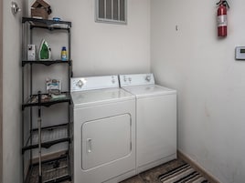 Un cuarto de lavandería con dos lavadoras y secadoras de carga frontal blancas, un estante metálico con productos de limpieza, un extintor y una rejilla de ventilación en la pared.