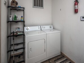 A laundry room with two white front-loading washers and dryers, a metal shelf with cleaning supplies, a fire extinguisher, and a vent on the wall. - Wine N Pines by Ruidoso Vacation Rentals (Alto)