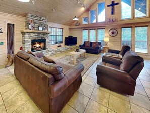 A cozy living room with a stone fireplace, leather sofas, a television, and large windows. - Wine N Pines by Ruidoso Vacation Rentals (Alto)