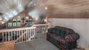 A cozy attic room with a wooden ceiling, a sofa, a desk, and a window overlooking greenery. - Wine N Pines by Ruidoso Vacation Rentals (Alto)
