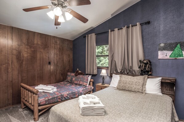 A bedroom with two beds, wooden headboards, a ceiling fan, a window with curtains, a nightstand with a lamp, and a wall painting. - Tis Holiday Haus by Ruidoso Vacation Rentals (Ruidoso)