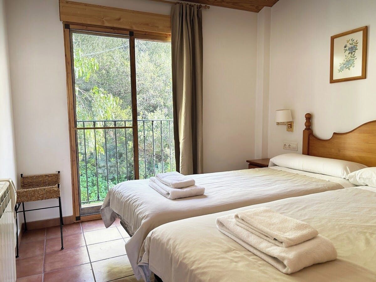 A hotel room with two beds, a view of greenery through a sliding glass door, a wooden headboard, and a small table with a lamp.