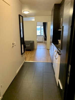 Um corredor estreito com uma porta à esquerda, um sofá cinzento ao centro e uma área de cozinha à direita.