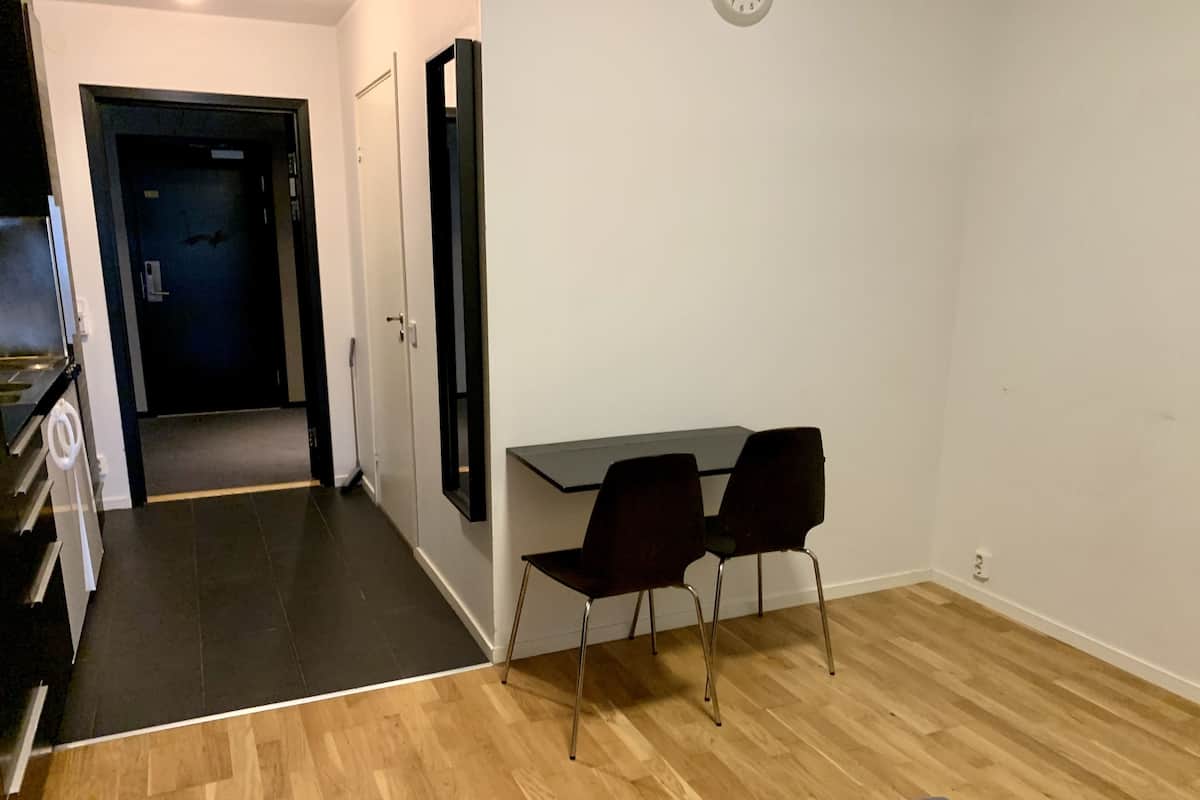 A room with a wooden floor, a dining table with two chairs, a black-framed mirror, and a door leading to another room.