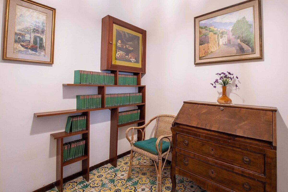 A room with a bookshelf, a wicker chair, a wooden dresser, and two framed paintings on the wall.