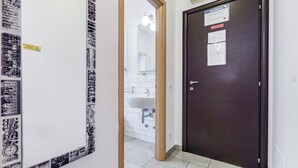En hall med en mörk trädörr, en vit vägg med en anslagstavla och en skymt av ett badrum med handfat och spegel.